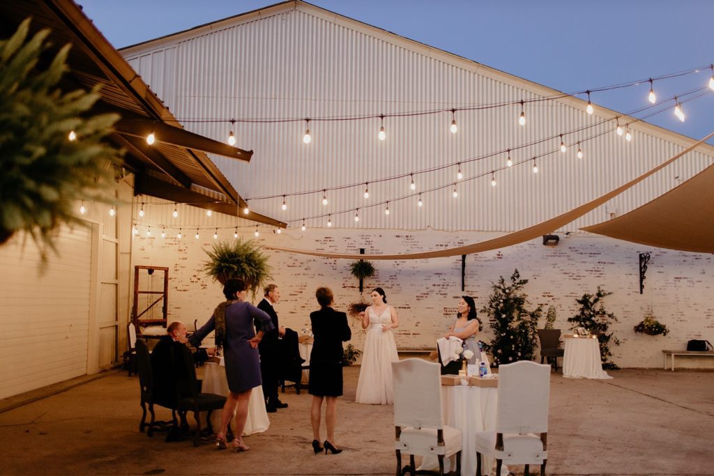 Outdoor Patio space on a summer evening. The patio space features string lights and sun shades, along with white washed brick and ferns/greenery. A Bride speaking with some of her guests are at the center of the image.