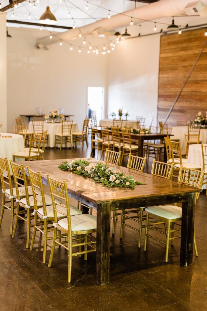 A interior shot of the Westside Warehouse Modern Industrial space. Highlighting a farm table used during a reception. The farm table is surrounded by gold chairs, and decorated with white flowers and greenery. String lights hang from the rafters providing a warm atmosphere.