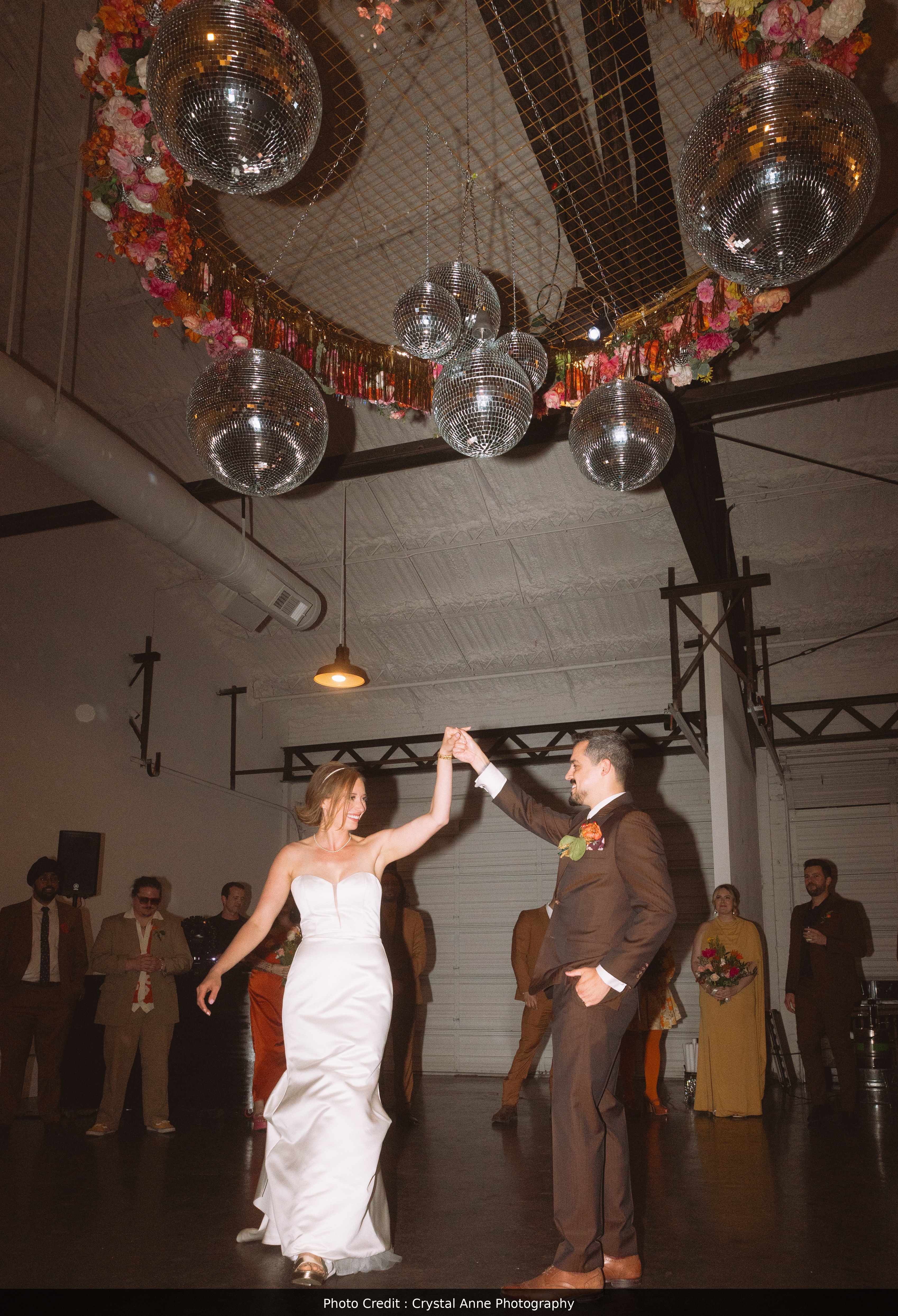 This image highlights a bride and groom enjoying their first dance, while surrounded by their wedding party. The image is set in a white modern industrial warehouse, and the dancing is under a decorative disco ball installation, hanging from the rafters. The bride and groom are smiling at one another and holding hands, the bride in a long white dress and the groom in a brown suit.