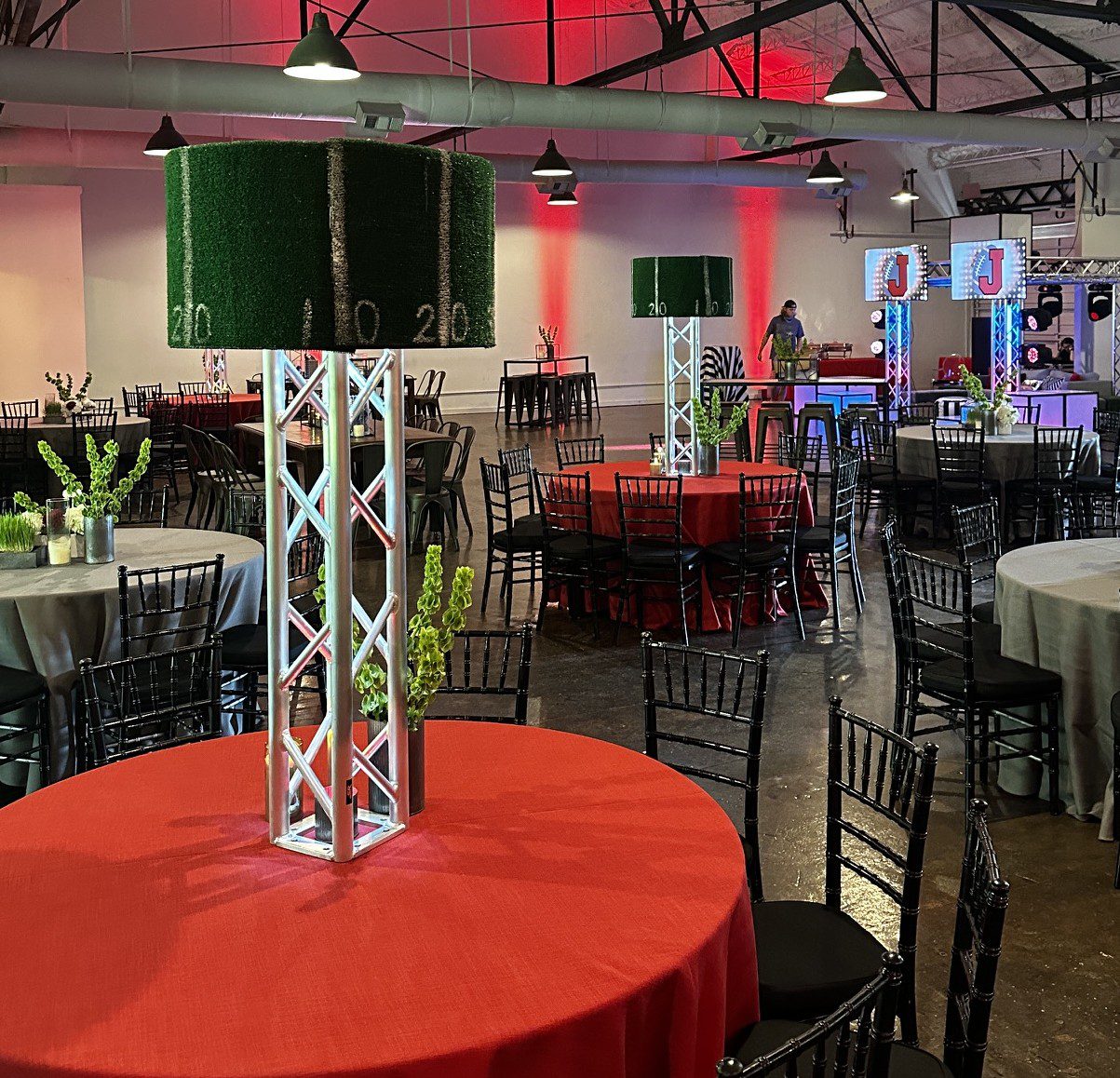 This image features an indoor party with lots of black chairs surrounded red and grey tables. The party has a clear sports theme, and an elevated DJ booth is visible in the background of the image. There is also red uplighting.