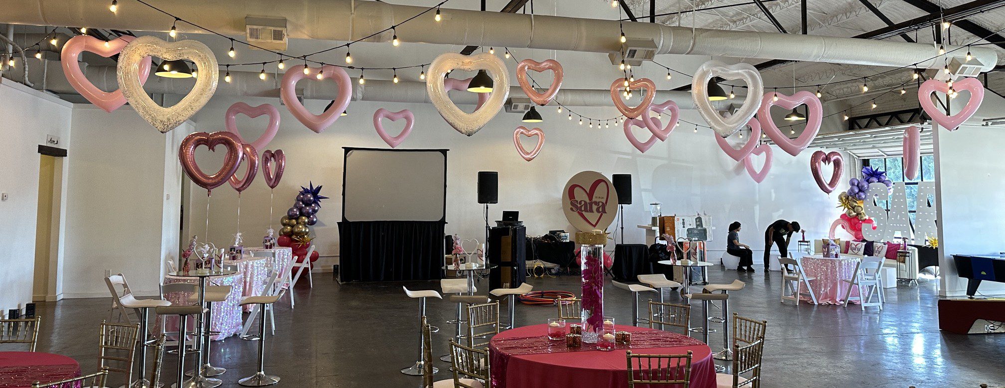 The image shows a modern industrial event space with string lights and heart balloons hanging from the rafters. There is an assortment of decor in the space, including red tables, gold chairs, a screen, live music, large letters, and video games. There is a lot of natural lighting coming in through the windows on the right side of the space.