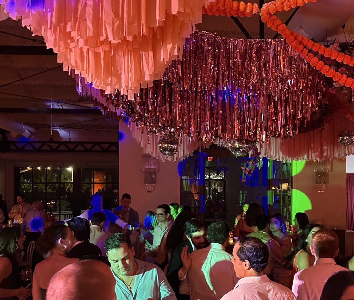 This image shows a crowded dance floor underneath a hanging decorative installation, featuring tassels and other reflective decorations. There are lots of colors reflecting on many surfaces, with guests dancing in the foreground and background.