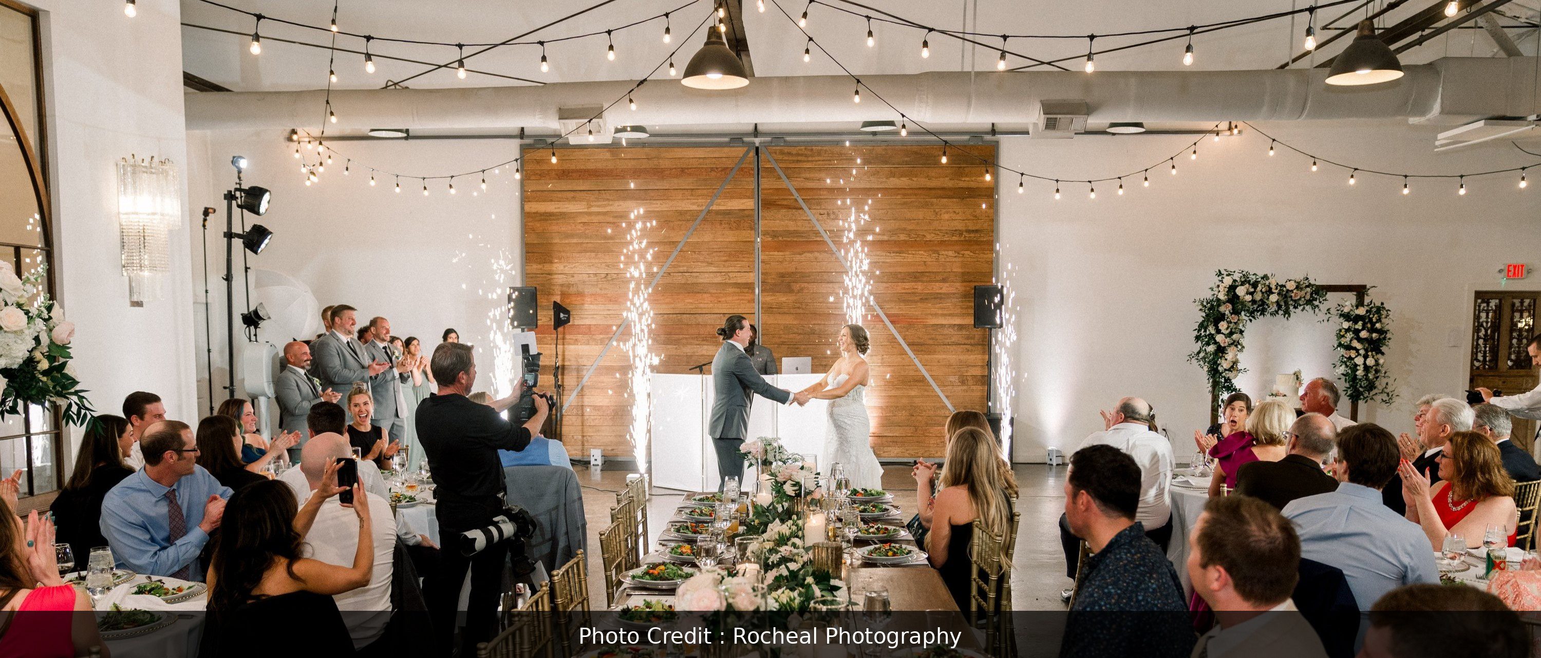 This image highlights a smiling bride and groom during their first dance. They are facing each other in the center of the room, with all their guests surrounding them and taking their picture. There are a set of large doors in the back drop, as well as a DJ setup. Cold sparklers are shinning in the background, illuminating the scene and providing a breathtaking atmosphere.