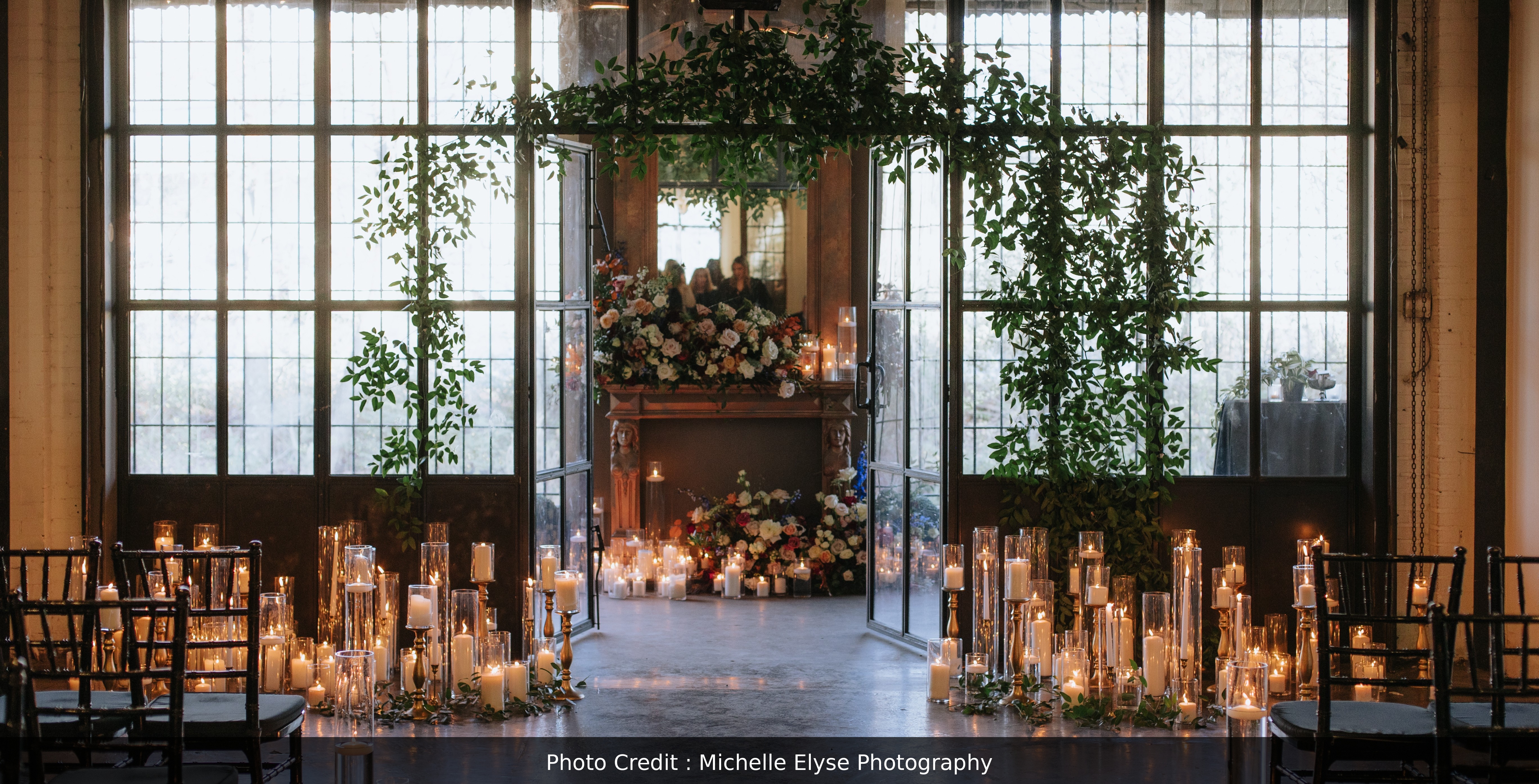 This image highlights a ceremony backdrop of a mantle and metal and glass doors. The doors are open, leading to the mantle. There are many lit candles set up in front of both the mantle and the doors. Lighting is shinning through the glass and doors. Some greenery is also installed around the opening, and there is a large floral arrangement both on top of the mantle, and inside it. Overall, the image has a warm, romantic, rustic, and whimsical look.